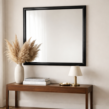 Decorative mirror in a black high-gloss frame hanging above a wooden console in a bright living room — styled with pampas grass and a brass table lamp